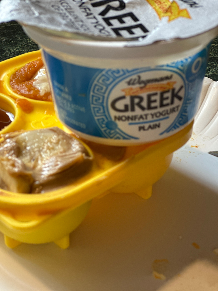 small tub of Greek yogurt sitting on top of yellow pupsicle tray with peanut butter
